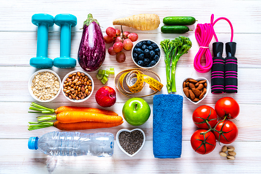 Exercising and vegan food concept: overhead view of dumbbells, jump rope, water bottle, towel, tape measure and healthy fresh multi colored organic vegetables, fruits, seeds and nuts arranged side by side on white background. The composition includes broccoli, eggplant, cucumber, celery, tomato, carrot, grape, apple, blueberry, chia seeds, pinto bean, almonds, pistachio, oat, among others. High resolution 42Mp studio digital capture taken with SONY A7rII and Zeiss Batis 40mm F2.0 CF lens