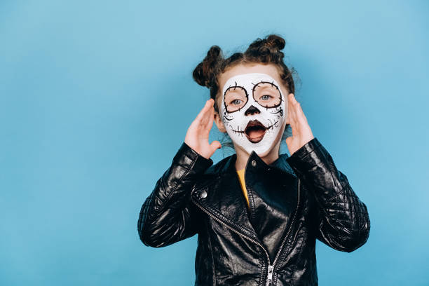 istock Stunned overjoyed little girl with bright scary makeup screams with excitement, keeps hands near mouth, has fun during halloween, has sugar skull on face, models over blue background with copy space 1276402861