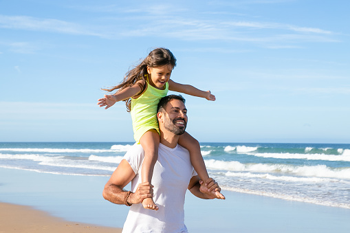 Joyful happy little daughter sitting on fathers neck and making flying hands. Excited dad and his girl walking on beach and having fun. Family outdoor activities concept