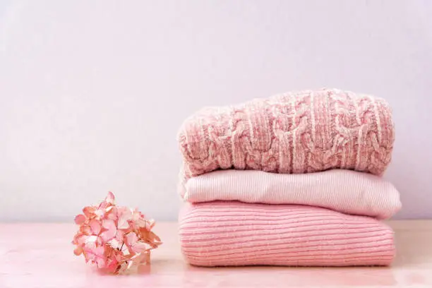 Photo of Stack of folded wool knitted sweaters in pink pastel colors laying on table for autumn winter season with flower