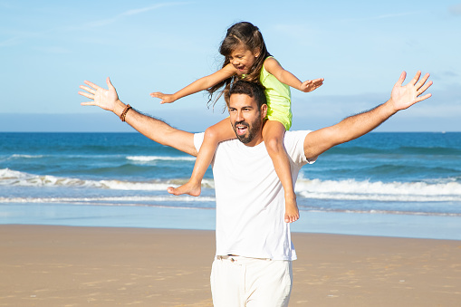 Excited dad and his girl walking on beach and having fun. Little daughter sitting on fathers neck and making flying hands. Family outdoor activities concept