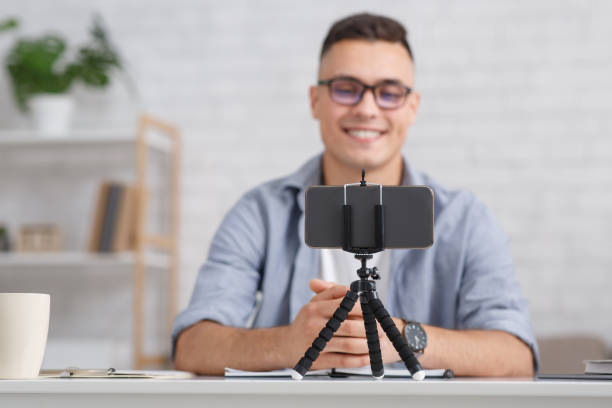 istock Modern hobby and remote work online. Focus on smartphone on tripod and smiling guy with glasses makes video for blog in interior of living room 1276408279