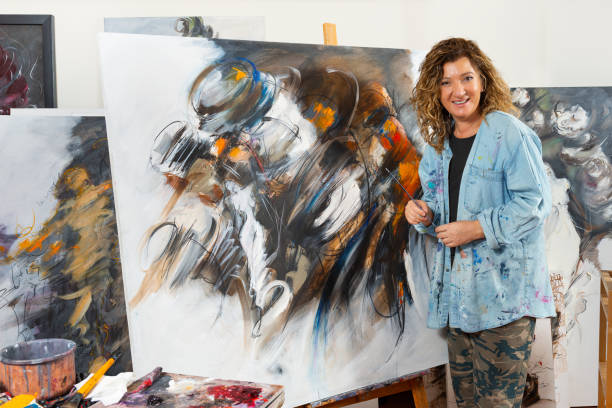 Portrait of fine art painter in her studio. She is standing in front of the canvas, looking at camera and smiling. Paint, brushes, painted canvases surrounds her.