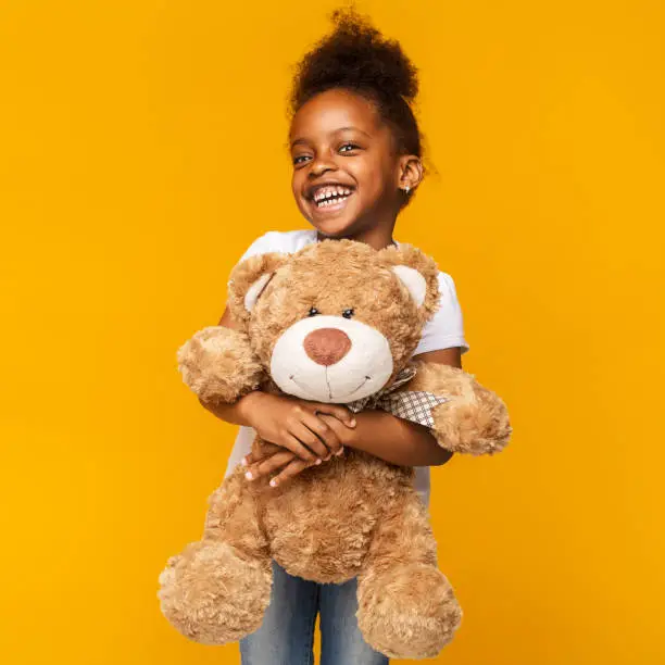Cute black little girl embracing big teddy bear and laughing over yellow background, kids toys concept