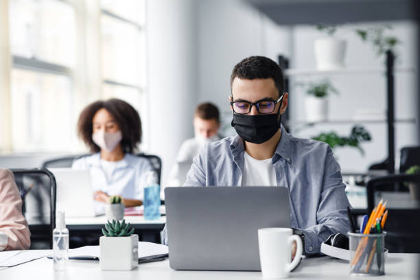 Office center workers are protected from virus outbreak during covid-19 epidemic. Young hipster man in glasses and protective mask works at laptop Office center workers are protected from virus outbreak during covid-19 epidemic. Young hipster man in glasses and protective mask works at laptop, with colleagues at workplaces in interior, free space illness prevention photos stock pictures, royalty-free photos & images