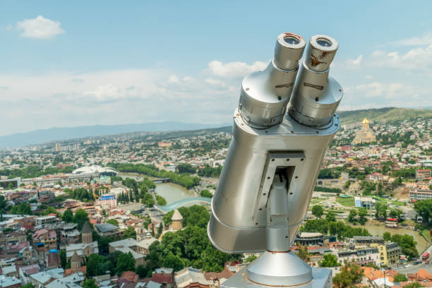 binocolo primo piano con vista su tbilisi, la capitale georgiana sullo sfondo in una giornata di sole. dispositivo grigio per visitare la città, visualizzazione stazionaria del binocolo, visualizzazione del binocolo con accettore di bolletta. - binoculars watching optical instrument closed foto e immagini stock