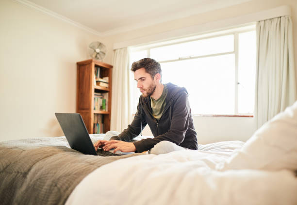 istock Young man sitting on his bed and working on a laptop 1276421704