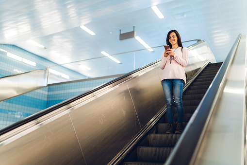 Young beautiful caucasian woman is checking social media or reading e-mails on smartphone on escalator at metro station.