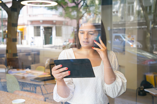 Serious businesswoman talking on cell, using tablet and looking at screen. View from street through window glass. Multitasking or communication concept