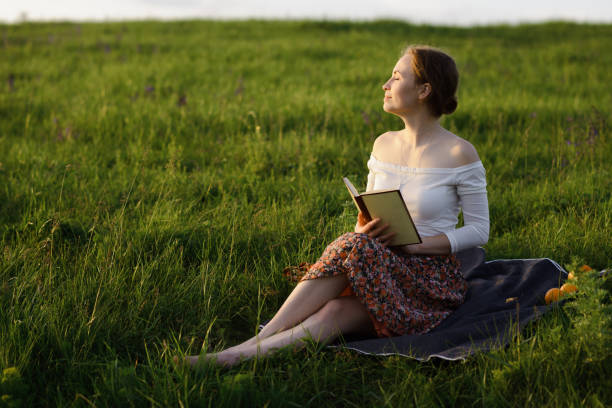 istock Young girl is reading a book and sitting on the grass while sun is setting down. Summer feeling 1276445192