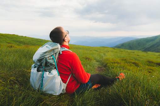 Man hiker with backpack feeling freedom and happiness resting at the beautiful meadow with mountain view during summer sunset - social distancing
