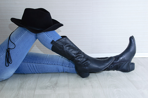 Woman legs in black cowboy boots with cowboy hat close-up. Sitting on a gray wooden background.