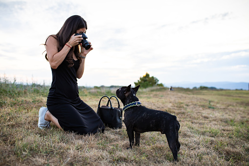 A young attractive woman is kneeling while taking a photo of her Frenchie in a park