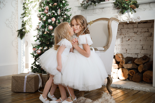 little beautiful girls in a white fluffy, elegant dress with magic wands sitting in a chair on the background of Christmas trees in the interior of the loft. new year holidays and vacations.