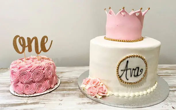 A pink smash cake and a white cake to eat on a table for a one year olds birthday celebration.