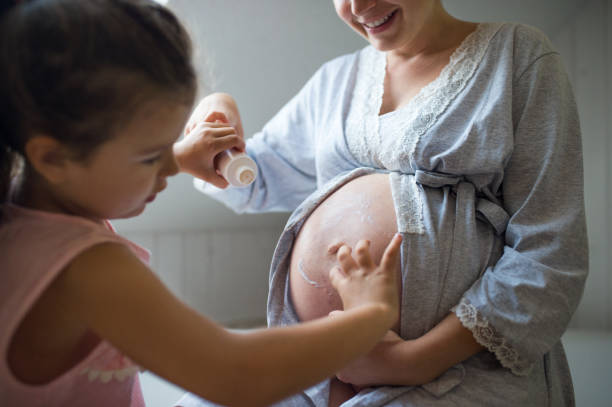 Small girl creaming belly of pregnant mother indoors in bathroom at home. Small girl creaming belly of unrecognizable pregnant mother indoors in bathroom at home. creaming stock pictures, royalty-free photos & images