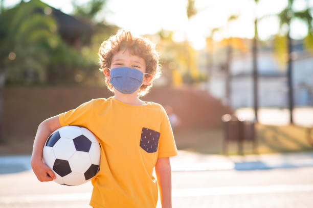 ребенок с маской защиты тканей лица с футбольным мячом на открытом воздухе во время пандемии - team sport enjoyment horizontal looking at camera стоковые фото и изображения
