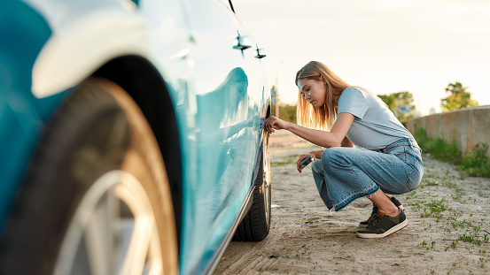 Attractive young woman checking air pressure of car tire on local road side while traveling, having troubles with her auto, checking wheel after car breakdown