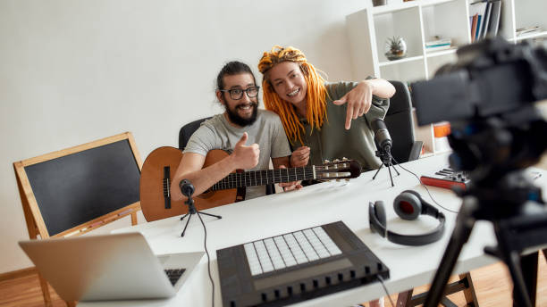 istock Man and woman showing thumbs up and pointing down, asking for subscription while recording video blog or vlog. Couple of musicians making music at home 1276485565