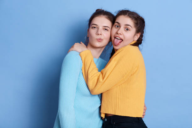 istock Two positive Caucasian young girlfriends hugging and having fun while posing isolated over blue background, ladies making grimace and showing tongue, wearing casual attires. 1276491454