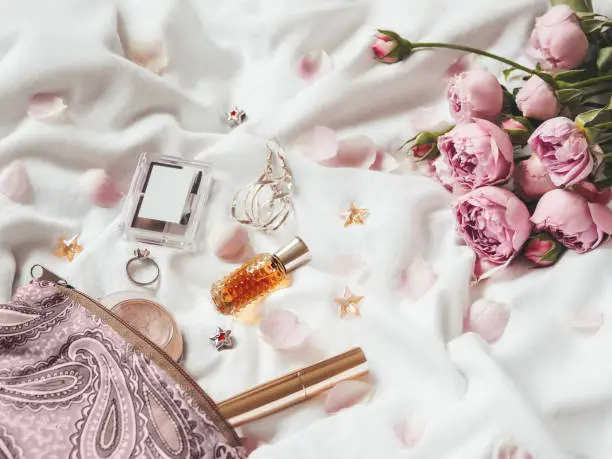 Photo of Cosmetic bag on crumpled white fabric background. Decorative cosmetic - perfume, mascara, eye and brow shadows. Cute and fashionable jewelry accessories. Pink roses and petals.