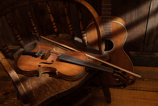 Western still life with a violin lying on a vintage saloon chair and a parlor guitar in the background. Sun streaks are raking across the barn wood wall onto the guitar and violin.