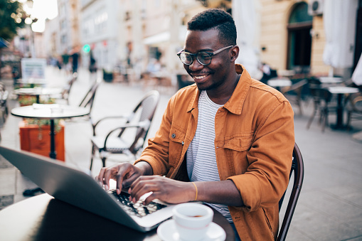 Happy young African American male freelancer sitting in a cafe, working online using his laptop and smiling