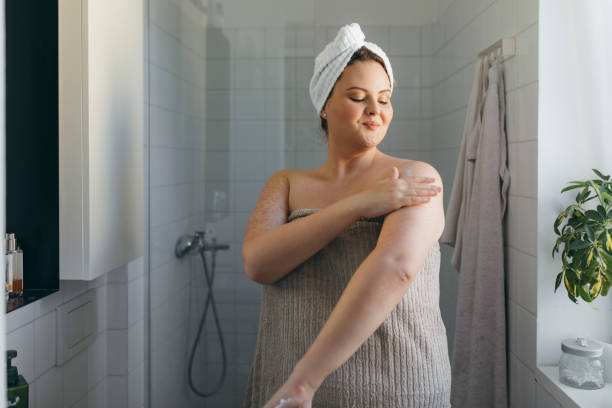 mooie overgewicht vrouw gewikkeld in een handdoek toepassing van body lotion na het hebben van een douche - douche stockfoto's en -beelden