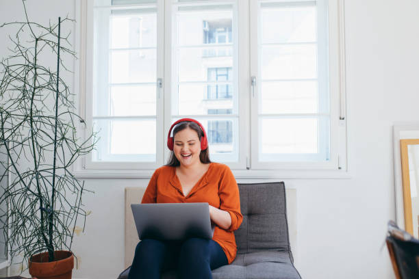 happy plus size mujer usando auriculares viendo algo divertido en su pc portátil - orange white audio fotografías e imágenes de stock