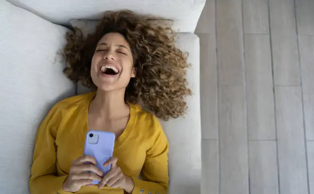 Happy woman laughing after watching something funny on her cell phone and lying on he sofa at home