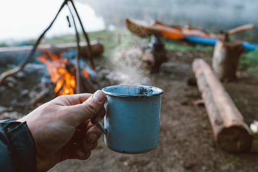 Photo of a male hand holding a iron mug in the background of the campfire and kayak boats