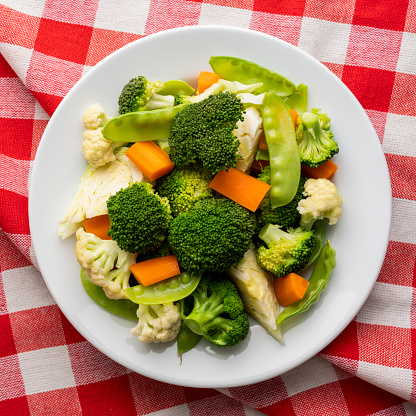 Colorful contrasting freshly steamed colorful greens consisting of broccoli, cabbage, carrots, sweet peas, and cauliflower on a red and white checked red gingham tablecloth background other healthy organic vegetables on a white ceramic plate sitting on an abstract white wood table background with lots of grain character, shot from directly above with good copy space on the left of the image.
