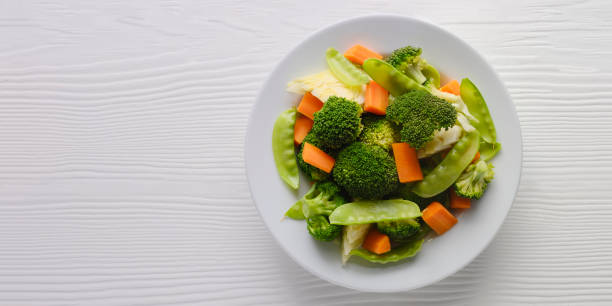 Fresh steamed broccoli, cabbage, carrots, sweet peas and cauliflower on a white ceramic plate on a white colored wood table background.