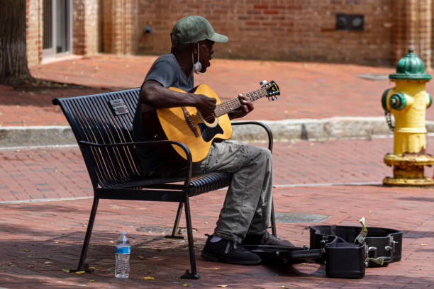 Annapolis, MD 08/21/2020: An elderly African American man wearing baseball hat and a face mask is playing classical guitar and singing while sitting on a bench on a street. He is a street musician.