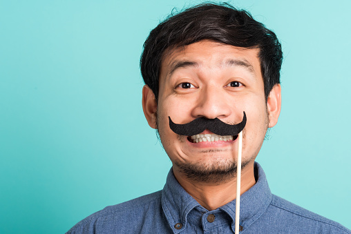 Portrait Asian happy handsome man posing he holding a funny mustache card or vintage fake moustaches on his mouth, studio shot isolated on blue background, Fathers day, November day concept