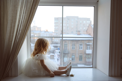 a little girl in a smart, fluffy white dress with a magic wand in her hands looks out the window and waits for Santa Claus or the tooth fairy. children's fairy tales and dreams. new year holiday.