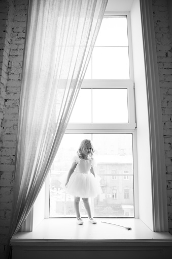 a little girl in a smart, fluffy white dress with a magic wand in her hands looks out the window and waits for Santa Claus or the tooth fairy. children's fairy tales and dreams. new year holiday.