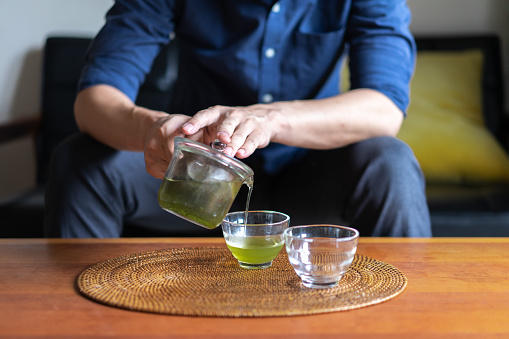 A man brewing Japanese tea in his living room at home.\nBrewing tea in a careful manner and savoring the tea slowly.\nSlowing down, relaxing time. Wellbeing. quality of life.