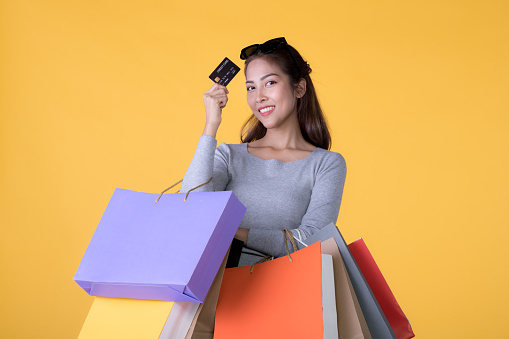 Beautiful young Asian woman with colorful shopping bags and credit card isolated on yellow background