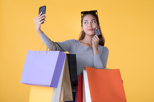 Beautiful young Asian woman with colorful shopping bags with a credit card and taking a selfie with smartphone isolated on yellow background