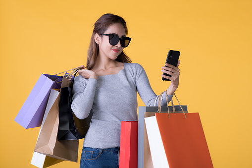 Beautiful young Asian woman with colorful shopping bags and smartphone isolated on yellow background