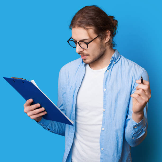 istock Bearded caucasian man with long hair making notes in a document holding a pen and folder on a blue studio wall 1276563882