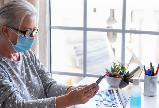 istock A concentrated senior woman with silver hair using laptop computer and smart phone working from home wearing face mask due to coronavirus, bright light from window- modern tech and social retired people 1276568428
