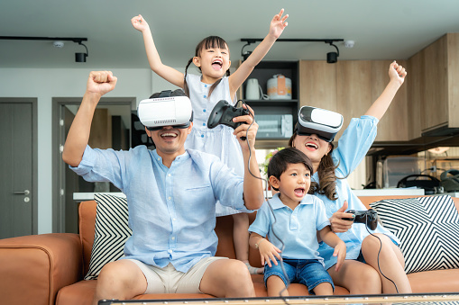 Happy Asian family at home on living room sofa having fun while they are looking mother and father playing games using virtual reality headset VR in living room at home.