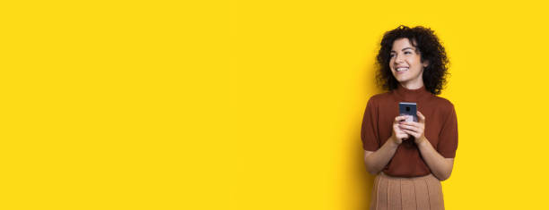 istock Curly haired brunette lady chatting on phone and smile at yellow studio wall with free space 1276577604