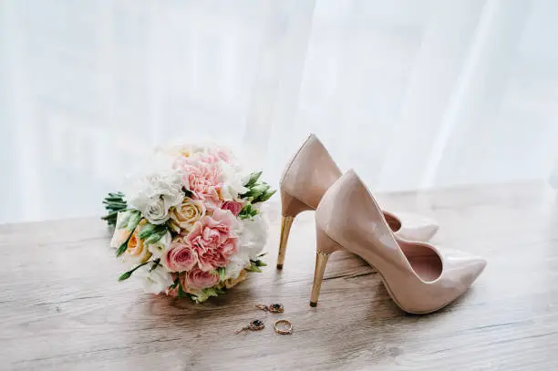 Photo of Wedding bouquet of the bride of pink flowers roses and greenery, stylish classic lacquered beige shoes, earring, and ring lying on wooden background. Bride accessories. Close up. Side view.