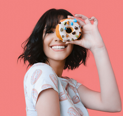 Caucasian brunette woman holding a tasty donut smiling at camera on a pink studio wall