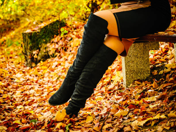 Woman in black leather boots is standing on asphalt pavement covered with orange oak leaves.