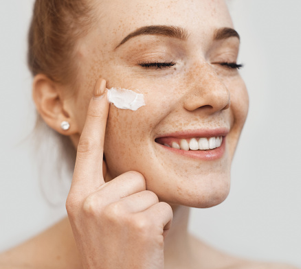 Freckled caucasian ginger lady applying on face anti aging cream and smile on a white studio wall