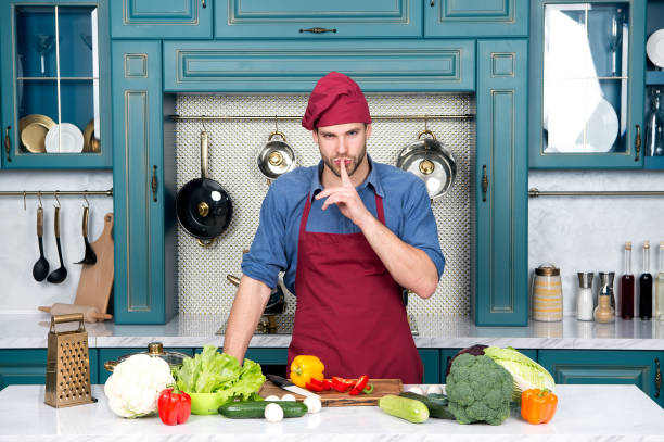 istock Chef with silence gesture finger at table with vegetables. Man cook secret dish in kitchen. Ingredients for cooking vegan food. Vegetarian menu and healthy diet. Food preparation and cooking recipes 1276586302
